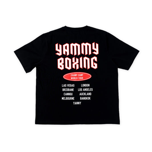 A black short-sleeve t-shirt shown flat against a white background, featuring a graphic print on the back with the words 'YAMMY BOXING' in large red and white bubble letters, a red oval badge reading 'CHAMP CAMP WORLD TOUR', and a list of travel destinations in white text.