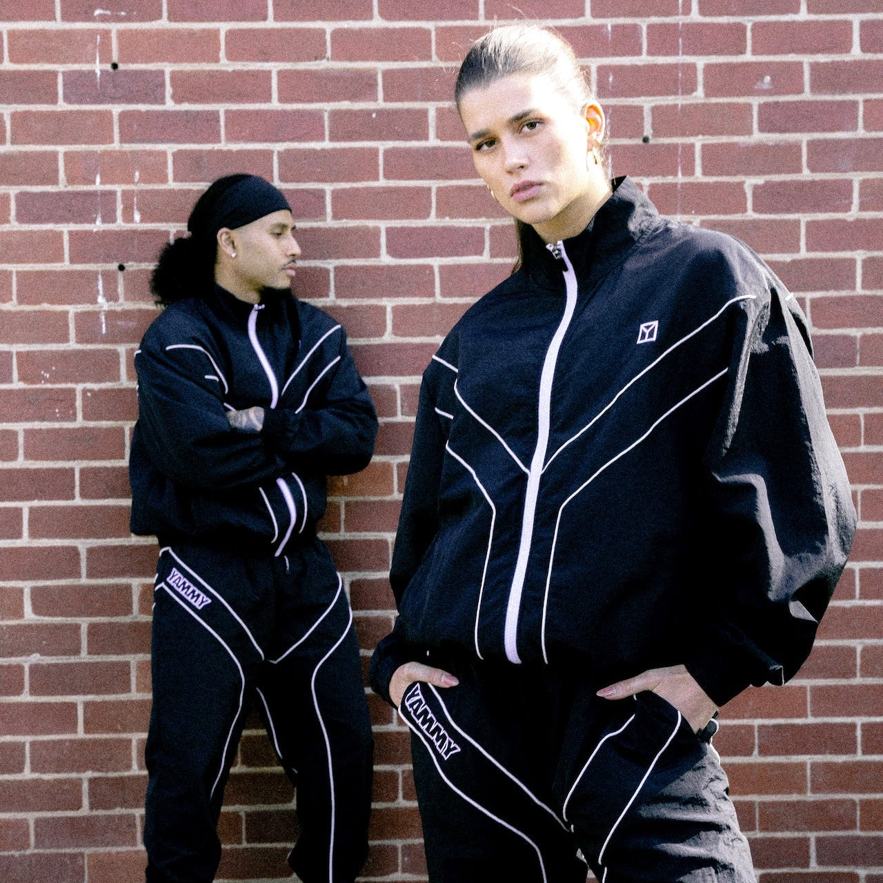 Two models wearing matching black tracksuits with white piping details, standing in front of a red brick wall.