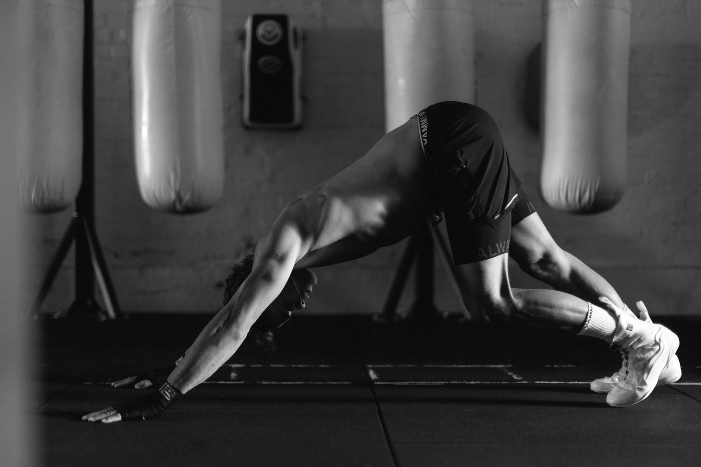 A black and white lifestyle photograph of a man performing a yoga stretch in a gym, wearing black athletic shorts and white sneakers, with hanging punching bags in the background.