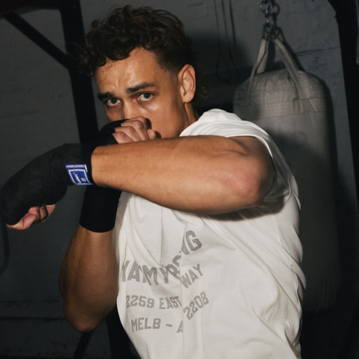 A man in a boxing stance wearing a white t-shirt with gray text, black hand wraps, and a black glove, set against a dark gym background with a hanging punching bag.