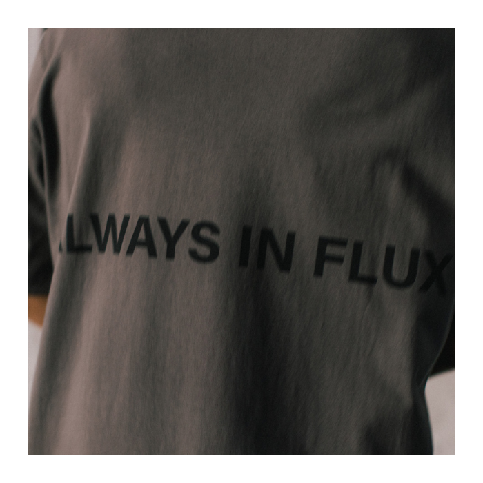 Close-up of a dark charcoal gray t-shirt featuring the text 'ALWAYS IN FLUX' printed in black across the chest.