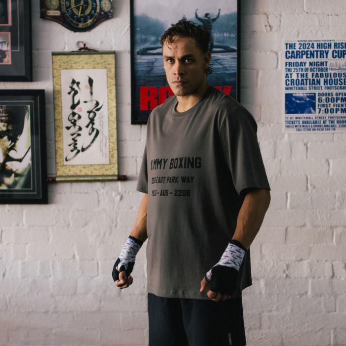 A man wearing YAMMY Boxing Parkway Charcoal T-Shirt, standing in a room with a white brick wall and boxing posters.