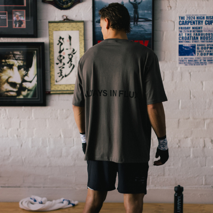 A man viewed from behind wearing a dark gray oversized t-shirt with 'ALWAYS IN FLUX' printed in black across the back, paired with black athletic shorts, standing in a room with framed wall art and a white brick wall.
