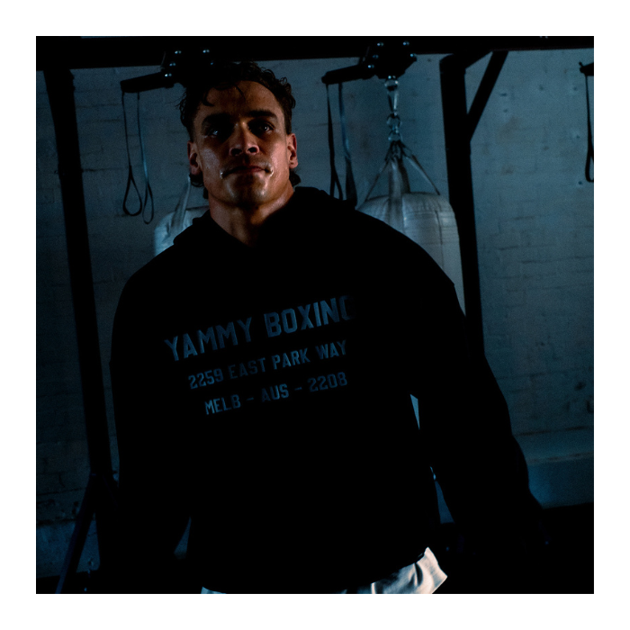 A man wearing a black hoodie with 'YAMMY BOXING' printed in blue on the chest, standing in a dimly lit boxing gym.