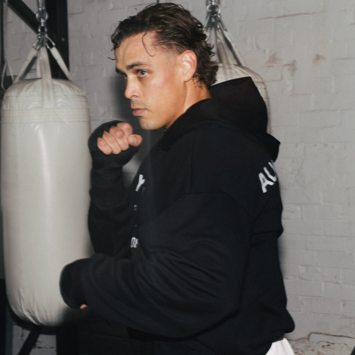 A man in a black hoodie and hand wraps poses in a gym in front of a white punching bag.
