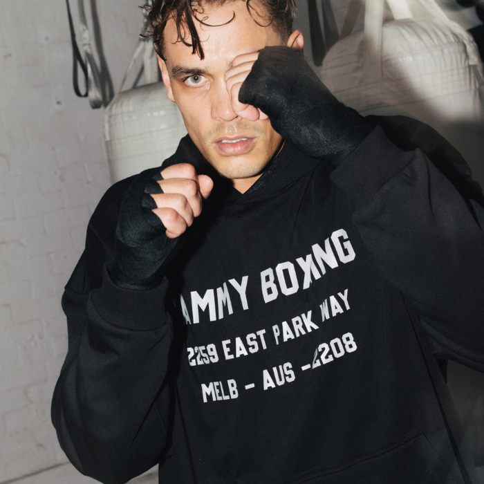 A man wearing a black YAMMY Boxing Parkway hoodie and black hand wraps poses in a boxing gym setting with white hanging bags in the background.