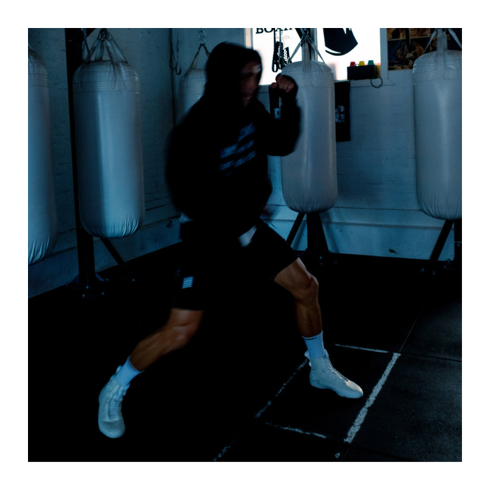 A person in a black hoodie and black shorts with white socks and white boxing shoes performing a punch in a boxing gym with hanging heavy bags.