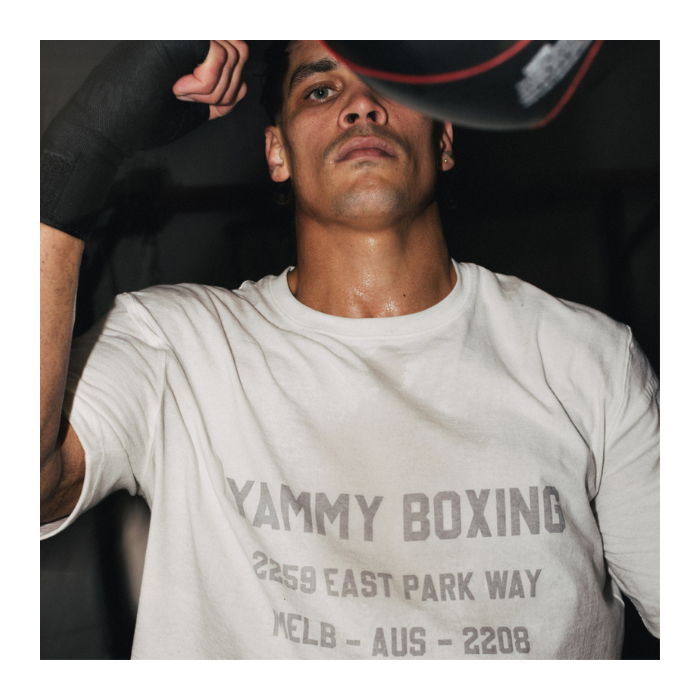 A man wearing a white graphic t-shirt with 'YAMMY BOXING' printed on the front, posing in a dark environment while holding a black cap.
