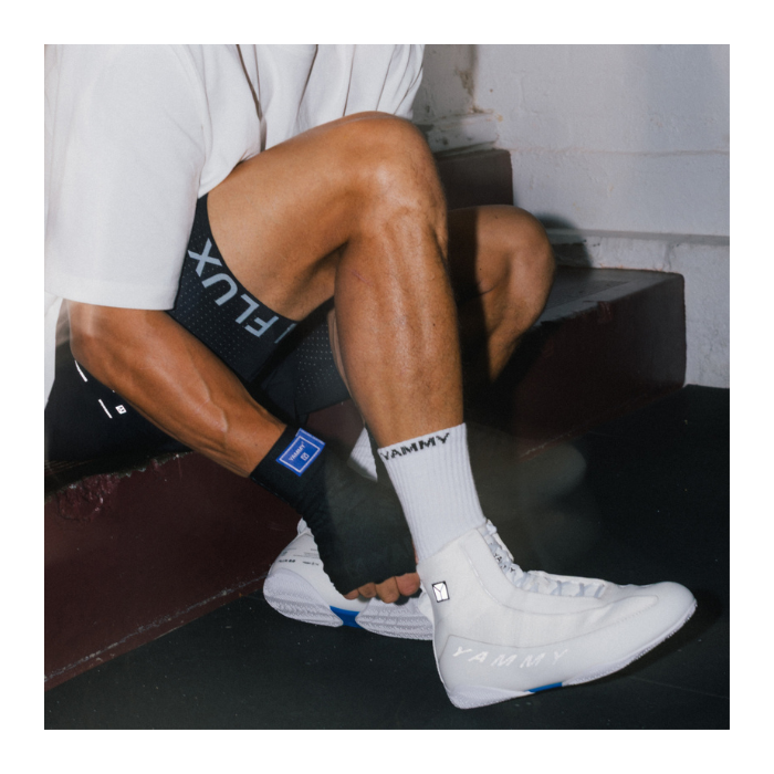A person sitting on dark steps wearing white YAMMY Boxing crew socks, white high-top sneakers, black shorts with white lettering, and a black wrist wrap.