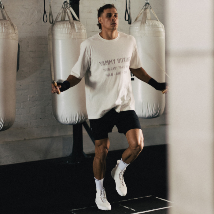 A man in a gym wearing a white graphic Parkway t-shirt with 'YAMMY BOXING' printed on the front, black athletic shorts, and white sneakers, jumping rope in front of hanging punching bags.