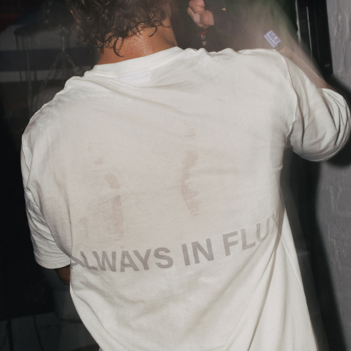 A person viewed from behind wearing a white long-sleeve t-shirt with the text 'ALWAYS IN FLUX' printed across the lower back in a dark gray tone.
