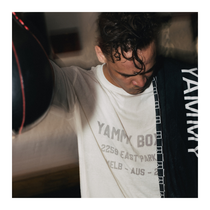 A man wearing a white graphic t-shirt with gray text and a black hoodie draped over his shoulder, standing next to a black punching bag in a dimly lit indoor setting.