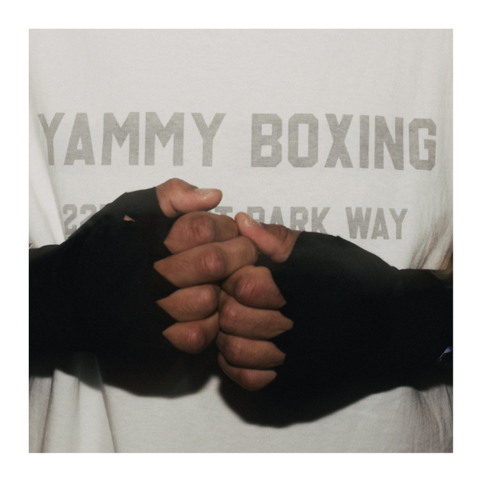 A close-up of two people's hands clasped in a fistbump, wearing black fingerless boxing gloves. They are wearing matching white t-shirts with 'YAMMY BOXING' printed in gray.