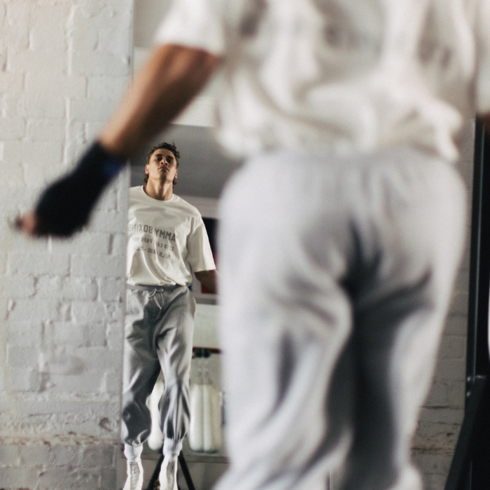 A man in a white graphic t-shirt and gray sweatpants stands in a gym, but the foreground is dominated by a heavily blurred figure wearing the same outfit, creating a shallow depth of field effect.
