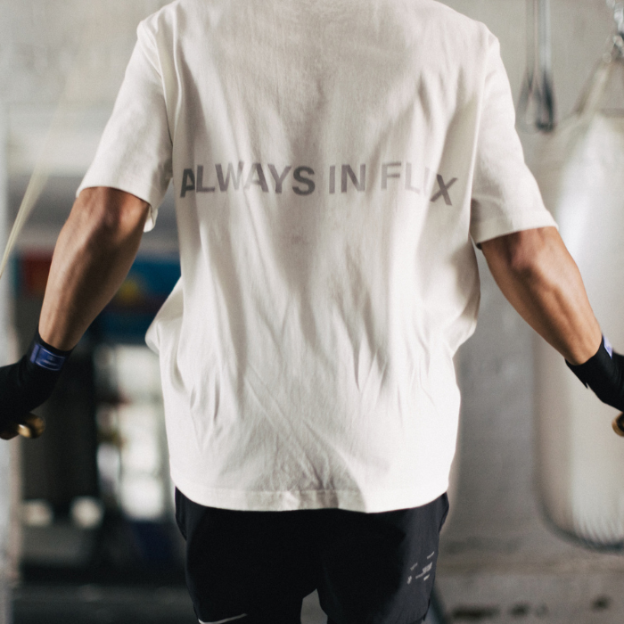 A person viewed from behind wearing a white short-sleeve T-shirt with the words 'ALWAYS IN FLUX' printed across the upper back in dark gray. The setting appears to be a gym with a punching bag in the background.