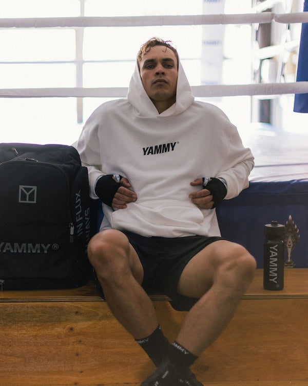 A man sits on a wooden bench inside a boxing ring wearing a white hoodie with 'YAMMY' printed on the chest, black shorts, and black hand wraps. A black YAMMY branded bag is on his left and a matching bottle on his right.
