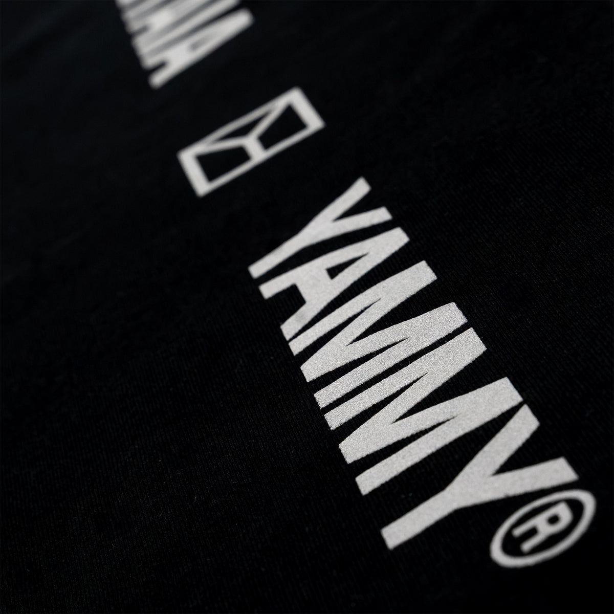Close-up of a black fabric surface with large white YAMMY branding and a small white geometric logo printed diagonally across it.