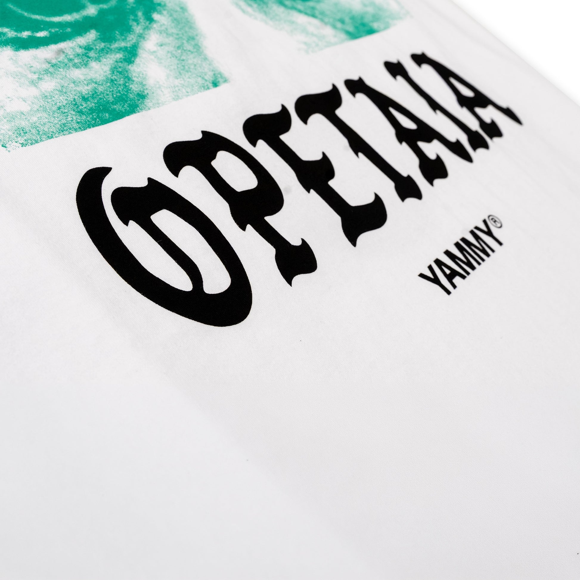 Close-up of a white graphic T-shirt featuring the word 'OPETAIA' in large black gothic-style lettering and 'YAMMY' in smaller black text, with a green abstract print visible at the top left.