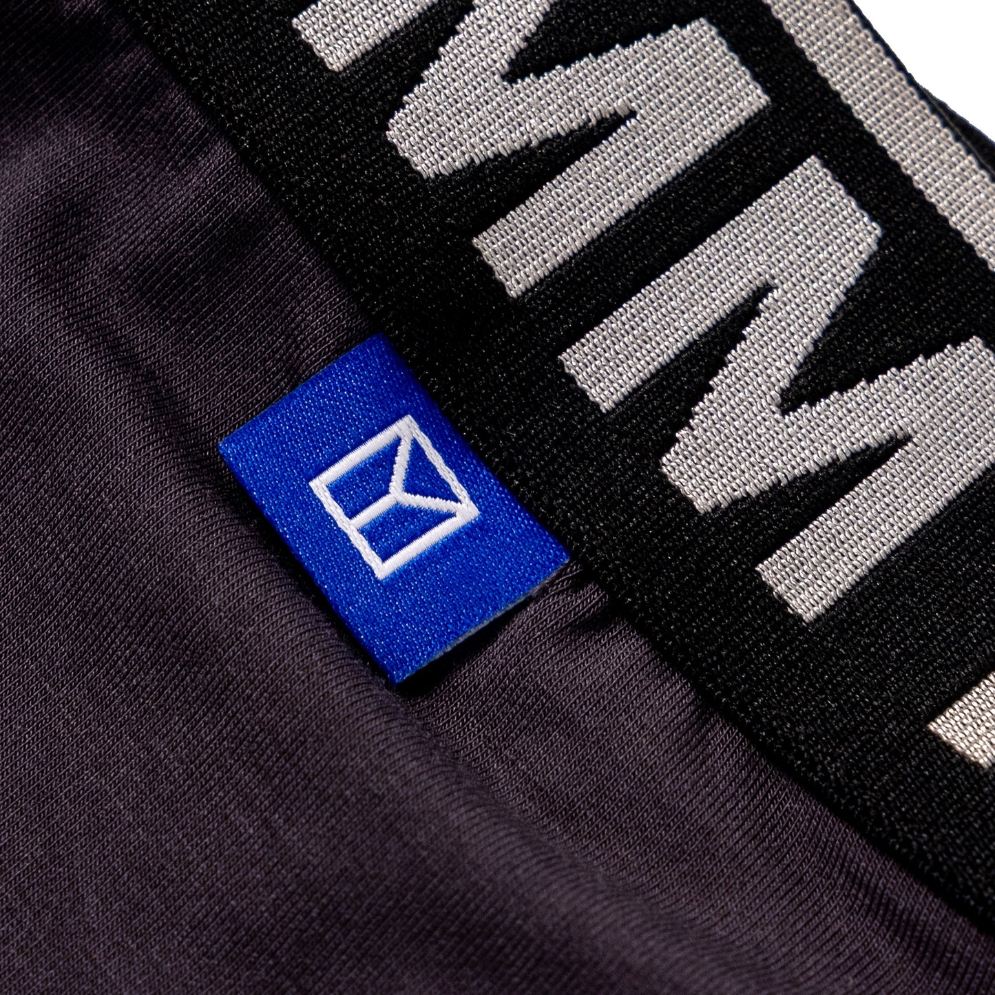 Close-up of a dark charcoal gray knit garment with a black elastic band featuring large white lettering and a small blue brand tag with a white logo.