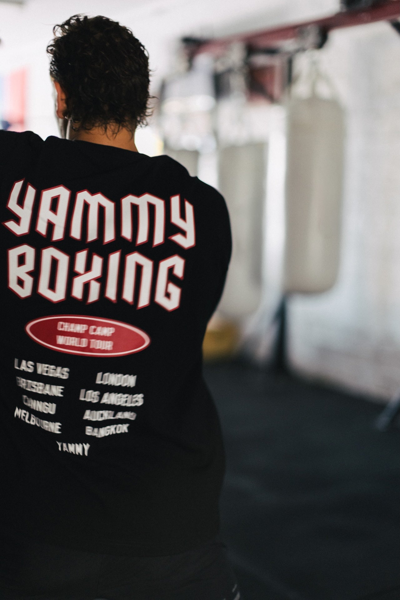 A person viewed from behind wearing a black long-sleeve hoodie with 'YAMMY BOXING' printed in large white letters with red outlines on the back, inside a boxing gym with hanging punching bags in the blurred background.