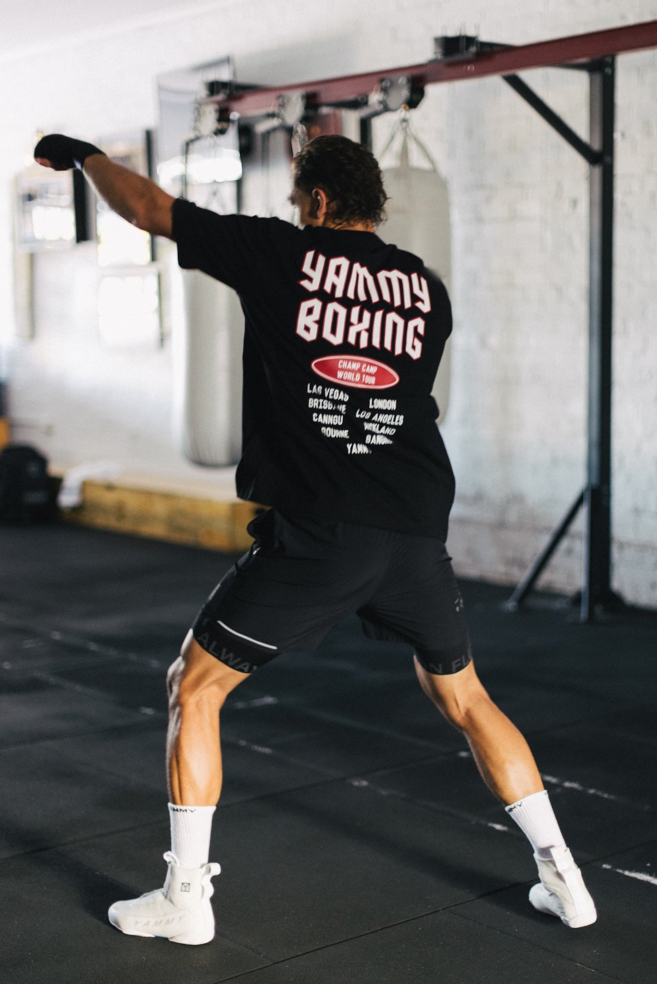 A man in a boxing stance wearing a black t-shirt with 'YAMMY BOXING' printed in white on the back, black boxing shorts, white socks, and white boxing shoes in a gym setting.