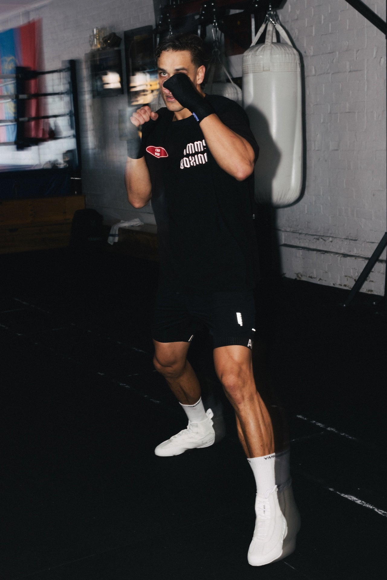 A man in a boxing stance wearing a black t-shirt with a red and white chest logo, black athletic shorts, white socks, and white boxing boots in a gym setting with a punching bag in the background.