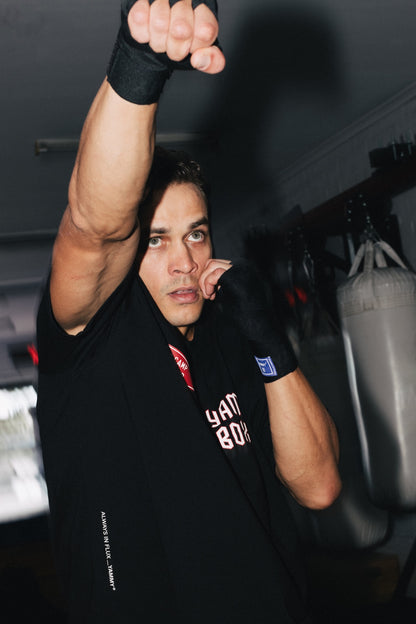 A man in a boxing stance wearing a black training shirt and black hand wraps, with a punching bag visible in the background of a gym.