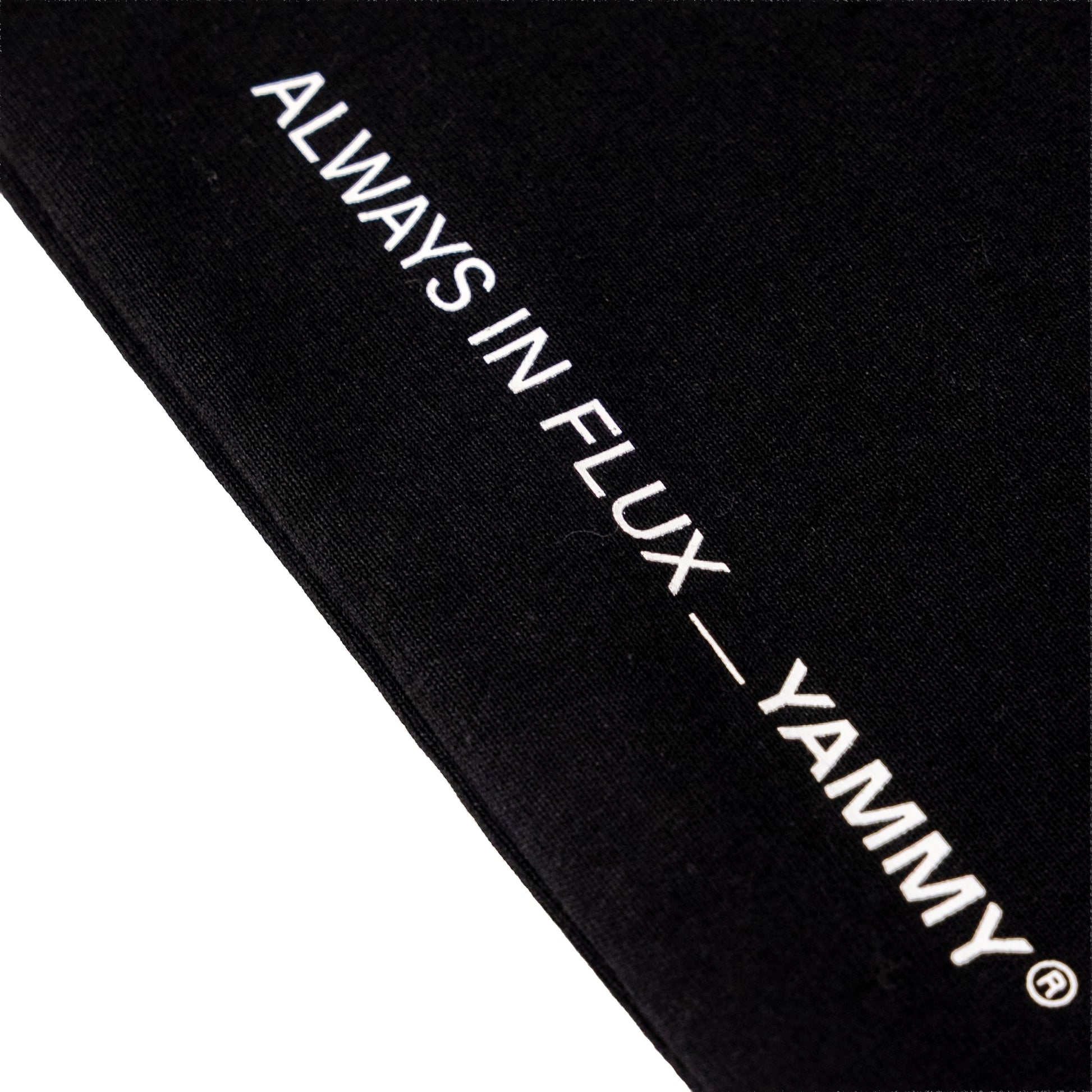 Close-up of a black fabric item with white embroidered text reading 'ALWAYS IN FLUX — YAMMY' on a white background.