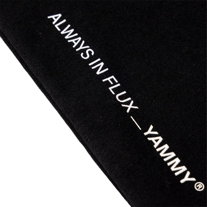 Close-up of a black fabric item with white embroidered text reading 'ALWAYS IN FLUX — YAMMY' on a white background.