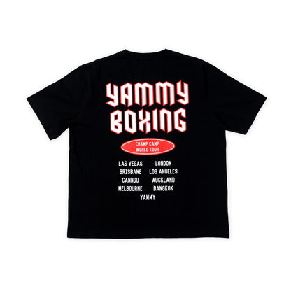 A black short-sleeve t-shirt shown flat against a white background, featuring a graphic print on the back with the words 'YAMMY BOXING' in large red and white bubble letters, a red oval badge reading 'CHAMP CAMP WORLD TOUR', and a list of travel destinations in white text.