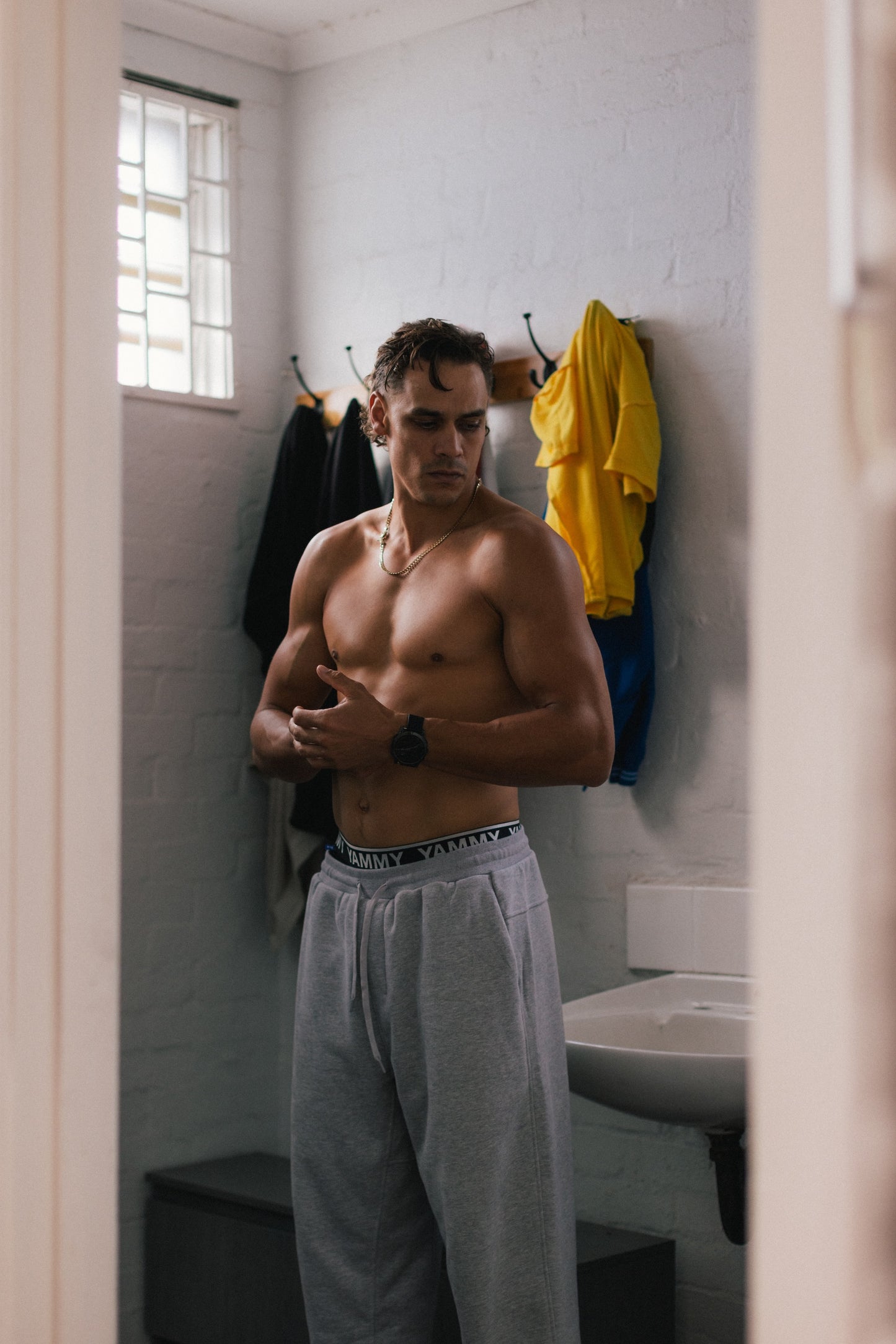 A shirtless man stands in a bathroom doorway wearing light gray sweatpants and a black waistband, with a sink, white brick wall, window, and hanging yellow shirt visible in the background.