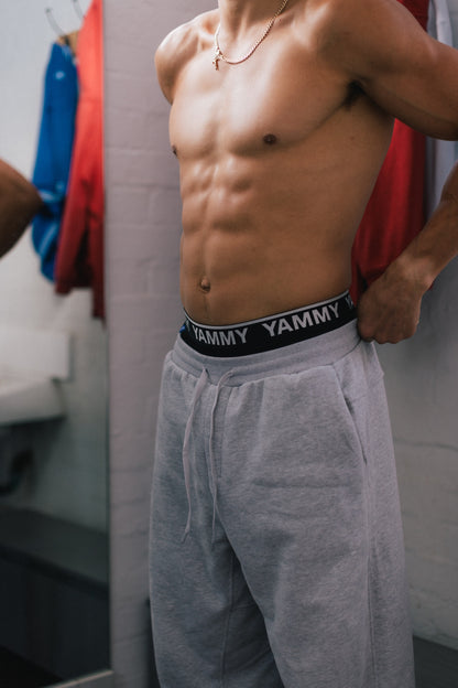 A shirtless man wearing light gray sweatpants and a black YAMMY brand waistband, standing in a fitting room or dressing area with clothing racks visible in the background.