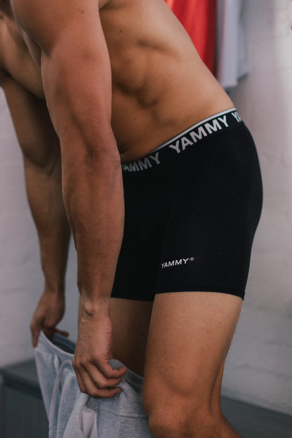 Close-up of a man wearing black Yammy branded boxer briefs, with the waistband text 'YAMMY' visible and a gray towel held in his hands against a light wall.