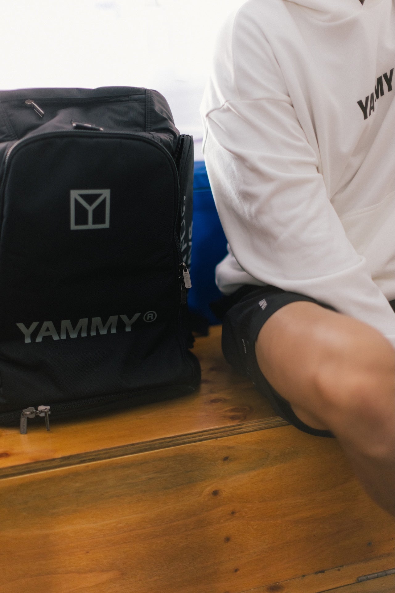A black YAMMY branded backpack sits on a wooden bench next to a person wearing a white sweatshirt and black shorts. The backpack features a silver geometric logo and the brand name 'YAMMY' printed in silver on the front.