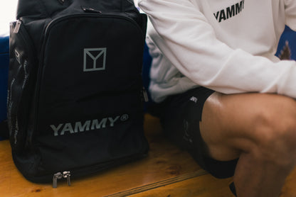 A black YAMMY branded backpack sits on a wooden bench next to a person wearing a white YAMMY long-sleeve shirt and black shorts.