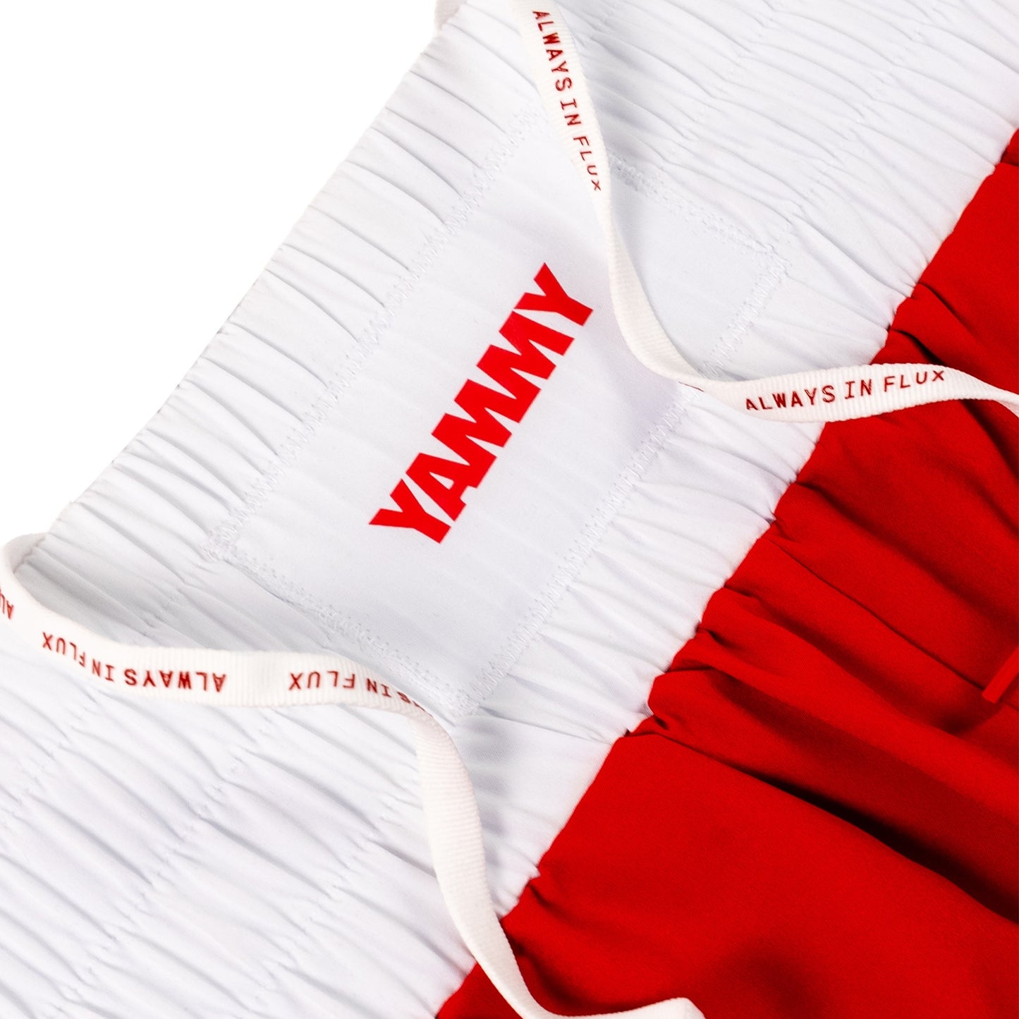 Close-up of athletic shorts with a white crinkled upper section, red side panels, and white drawstrings printed with the brand name YAMMY and the phrase ALWAYS IN FLUX.