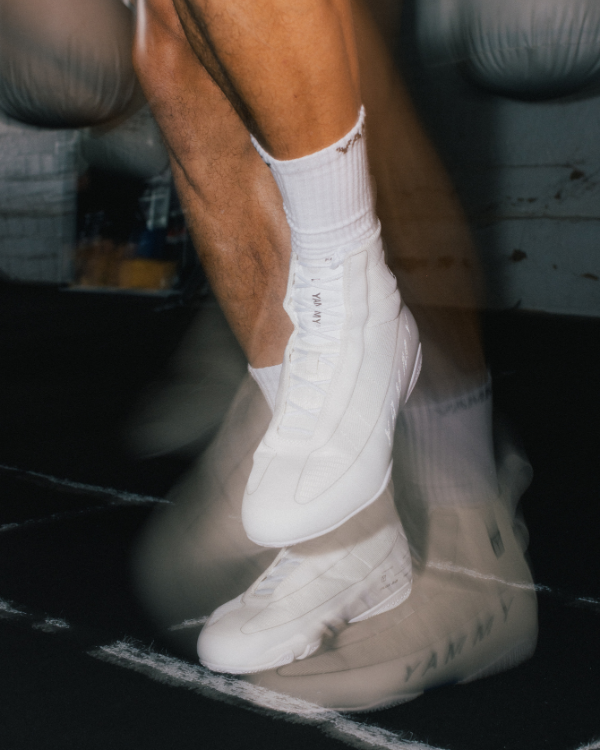 A pair of white high-top sneakers shown from different angles on a dark reflective surface. The shoes feature a minimalist design with tonal branding and small blue accents near the sole.