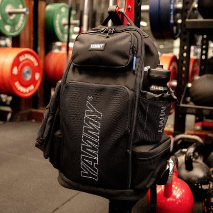 A black YAMMY backpack hanging on a gym rack, with weight plates and kettlebells visible in the background.