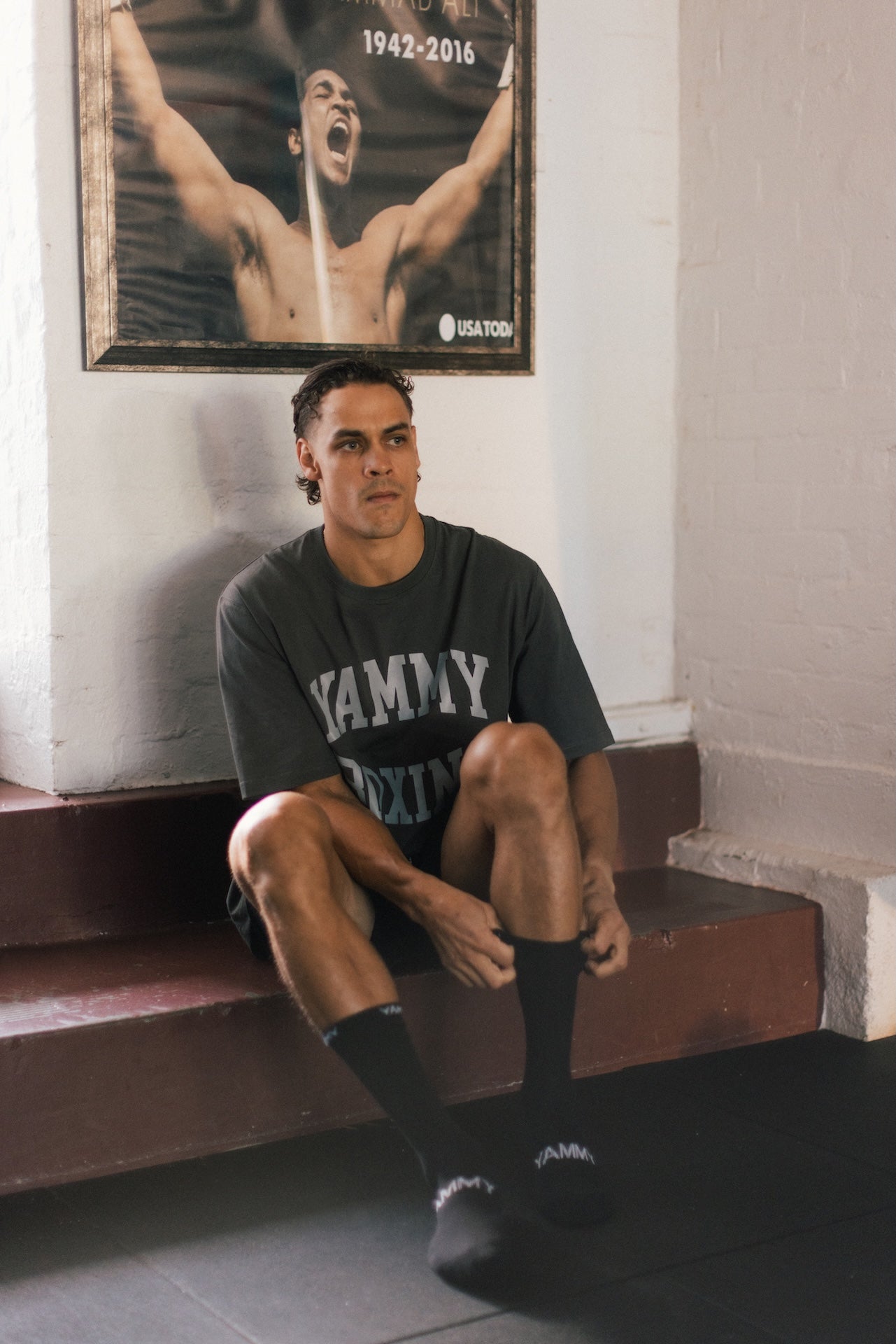 A man sits on wooden steps in a gym, wearing a dark gray graphic t-shirt with 'YAMMY Boxing' printed in white, black crew socks. A framed poster hangs on the white brick wall behind him.
