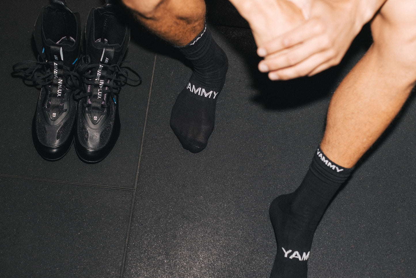 Black athletic socks with white 'YAMMY' branding worn by a person sitting on a dark gym floor, next to a pair of black sneakers.