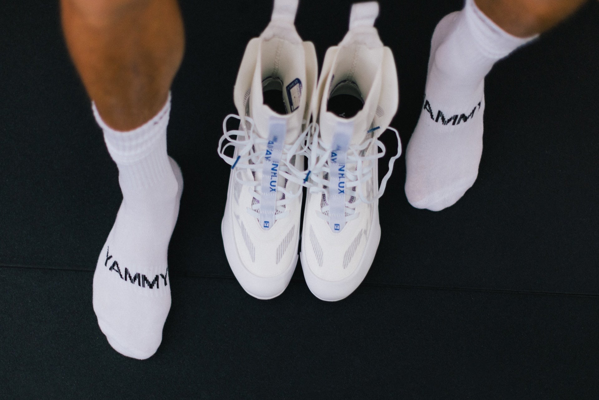 A pair of white high-top boots placed on a black surface, with white socks branded 'YAMMY' worn by a person whose legs are visible at the top corners of the frame.