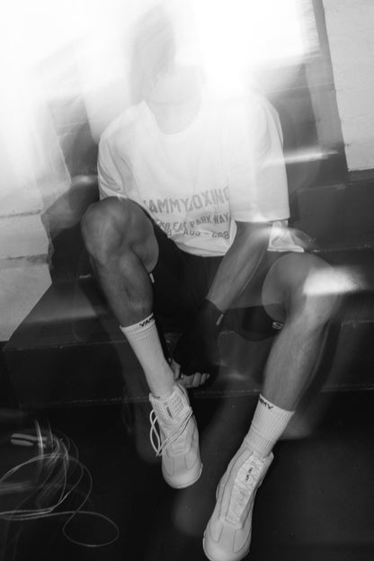 A person sitting on dark steps in a black-and-white photo, wearing a white graphic T-shirt, dark shorts, white socks, and white high-top sneakers.