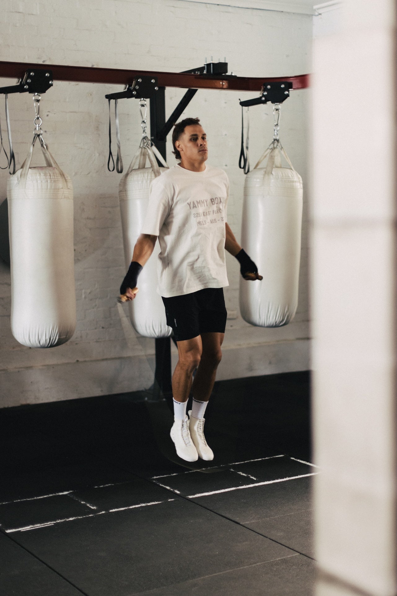 A man in a white graphic t-shirt, black shorts, and white sneakers jumps rope in a gym, with three white hanging punching bags suspended from a wooden overhead rig behind him.