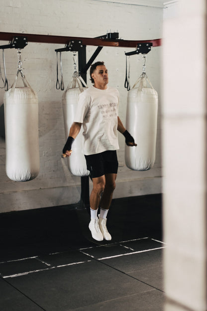 A man in a white graphic t-shirt, black shorts, and white sneakers jumps rope in a gym, with three white hanging punching bags suspended from a wooden overhead rig behind him.
