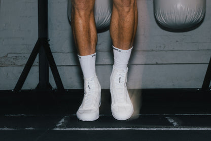 A close-up of a person's legs wearing white athletic socks and white high-top sneakers, standing on a black gym floor in front of a gray wall with hanging weights.