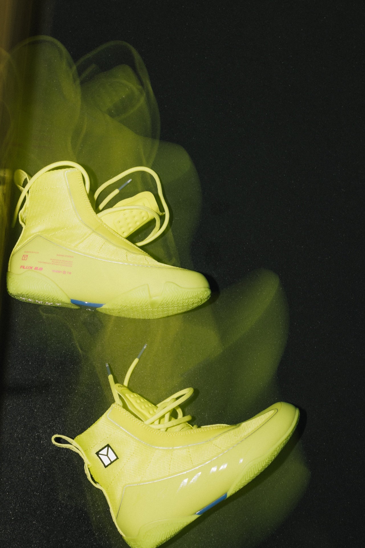 A pair of axis green boot displayed on a glossy black surface with motion-blurry reflections. The shoes feature a bright fluorescent yellow color, matching yellow laces, and small blue and pink graphic accents on the sides.