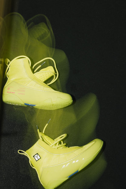 A pair of axis green boot displayed on a glossy black surface with motion-blurry reflections. The shoes feature a bright fluorescent yellow color, matching yellow laces, and small blue and pink graphic accents on the sides.