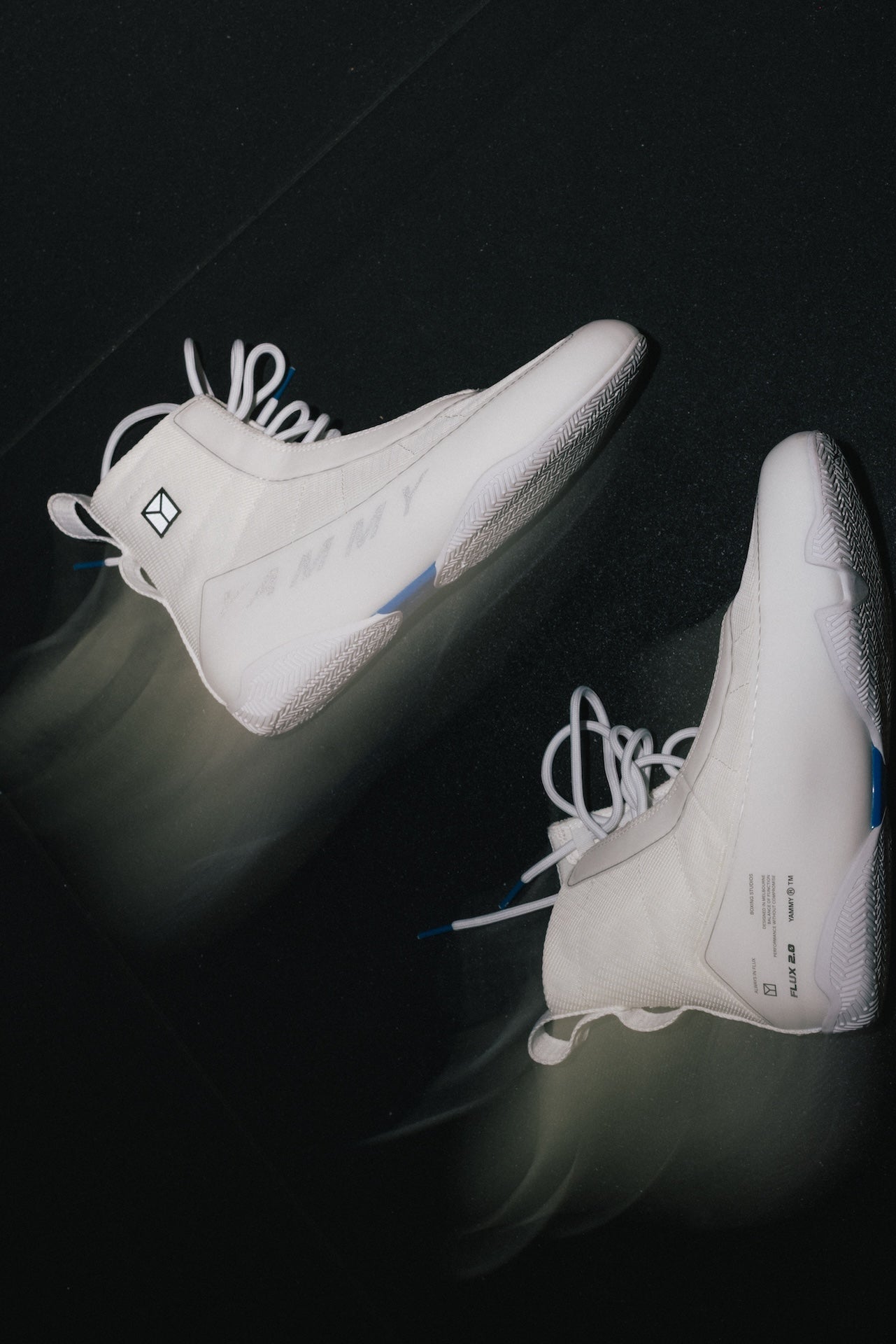 A pair of white high-top sneakers photographed from above on a dark reflective surface. The shoes have white laces, subtle blue accents near the midfoot and outsole, and small black logo details.