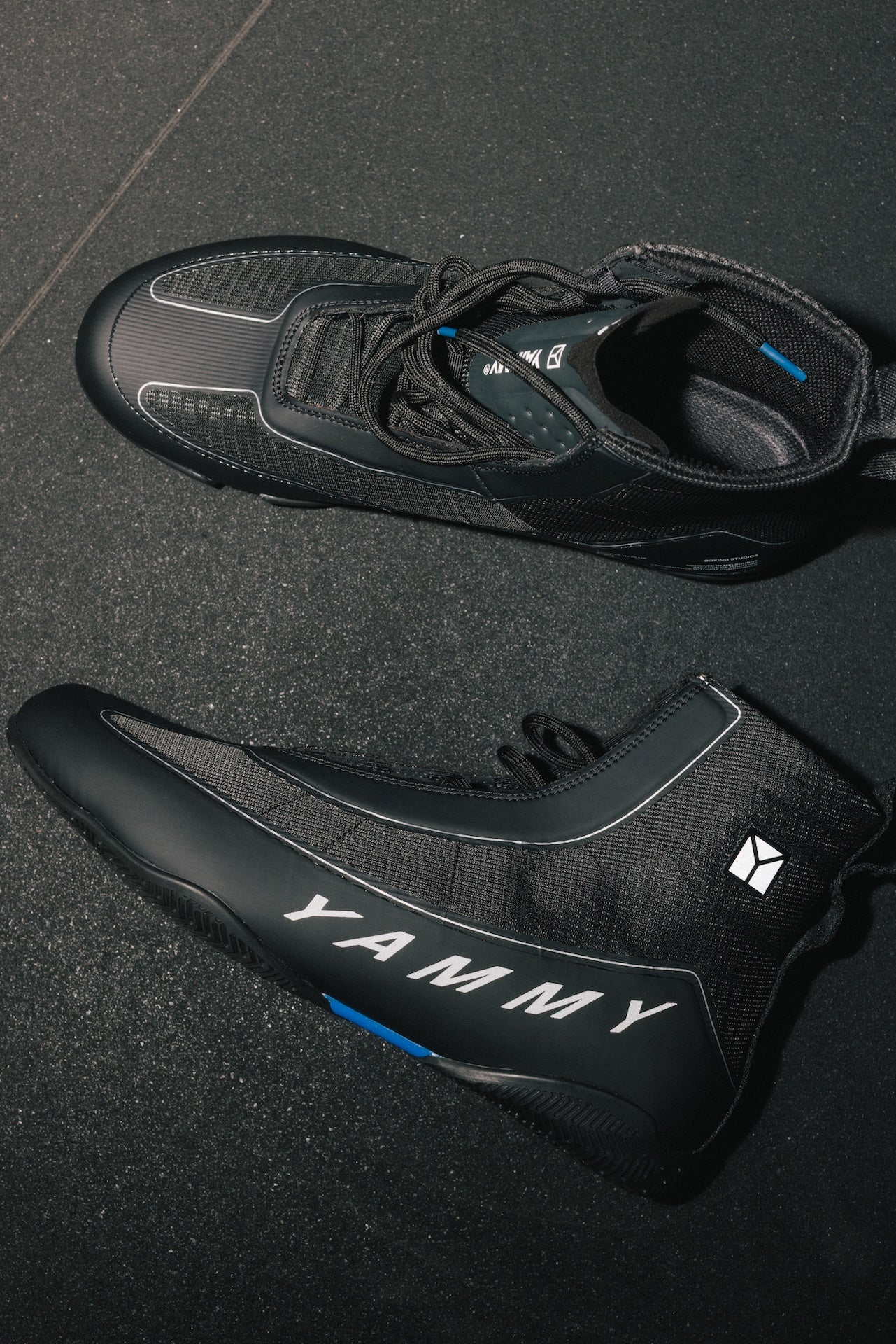 A pair of black high-top cycling shoes with white 'YAMMY' branding and small blue accents, displayed on a dark textured floor.