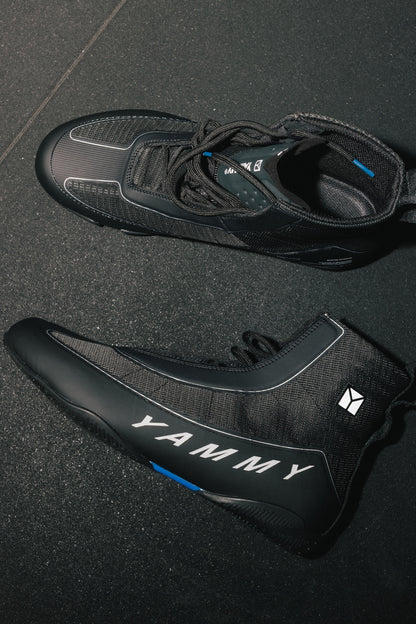A pair of black high-top cycling shoes with white 'YAMMY' branding and small blue accents, displayed on a dark textured floor.